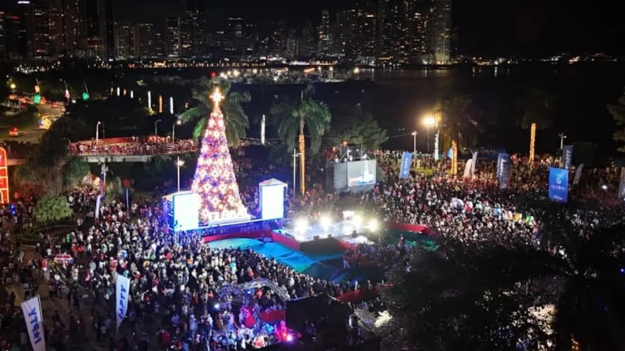 Multitud reunida de noche frente a un gran árbol de Navidad iluminado en la costanera con edificios al fondo.