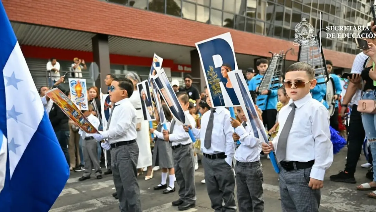 Niños con camisa blanca y corbata sostienen pancartas con letras durante un desfile escolar frente a una multitud.