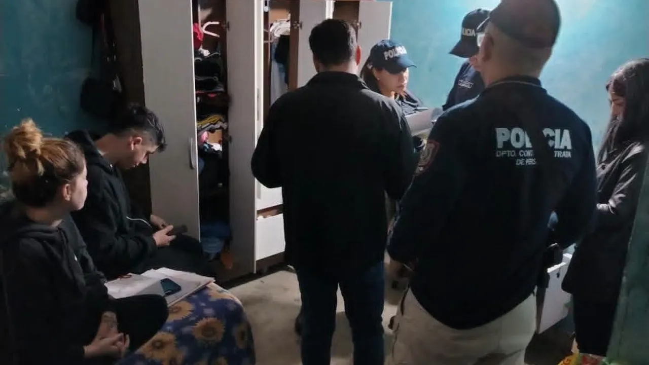 Agentes policiales y una fiscal inspeccionan una habitación y revisan documentos frente a dos jóvenes sentados.