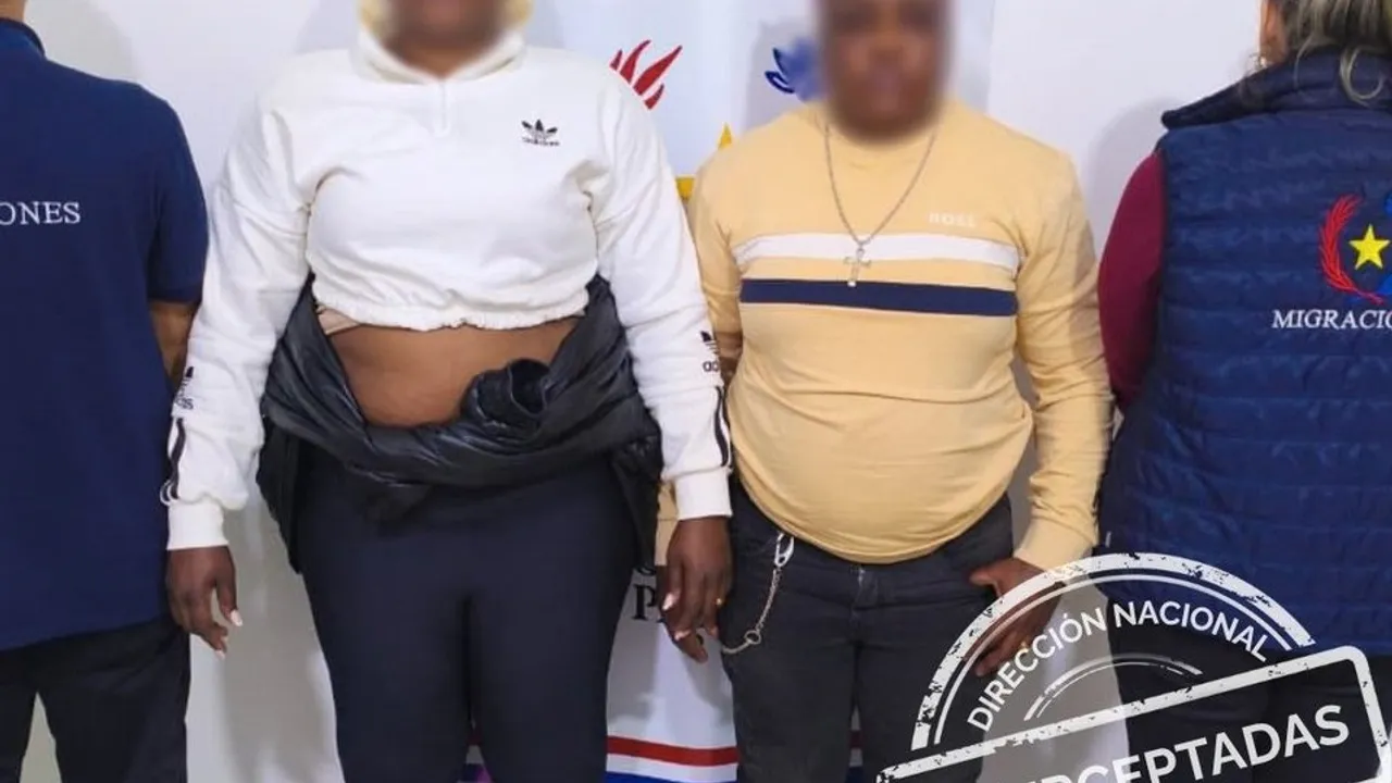 Dos mujeres con rostros difuminados permanecen de pie custodiadas por agentes con chalecos de Migraciones.