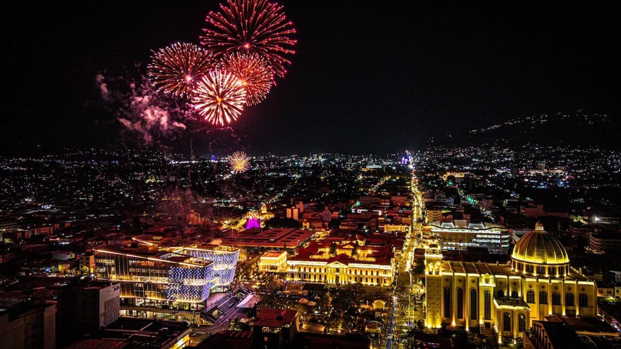Fuegos artificiales sobre edificios iluminados y la catedral del centro histórico de San Salvador de noche.
