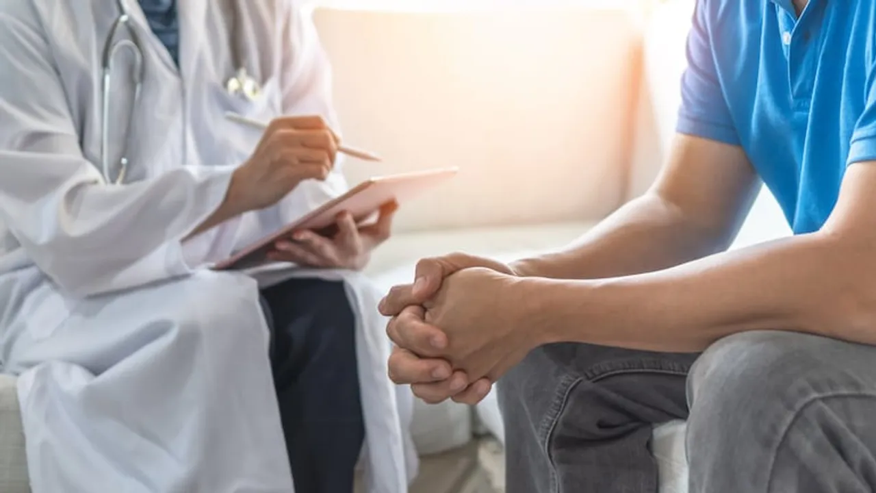 Médico con bata blanca y estetoscopio sostiene una libreta frente a un paciente sentado en el consultorio.
