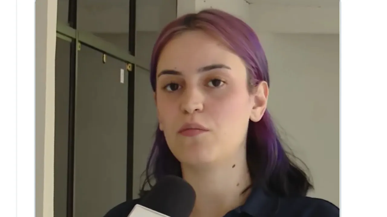 Joven con el cabello teñido de violeta brinda declaraciones frente a un micrófono institucional en un interior.