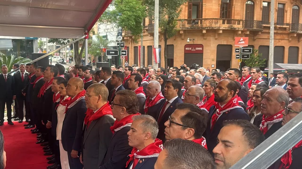 Dirigentes políticos con pañuelos rojos en el cuello participan de un acto partidario frente a una calle urbana.