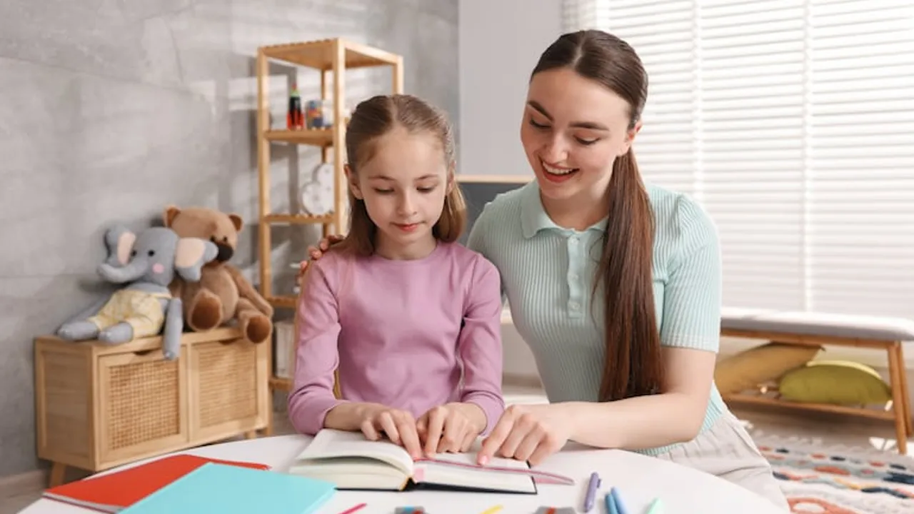 Niña sentada junto a una mujer mientras leen un libro con una regla sobre una mesa infantil con útiles