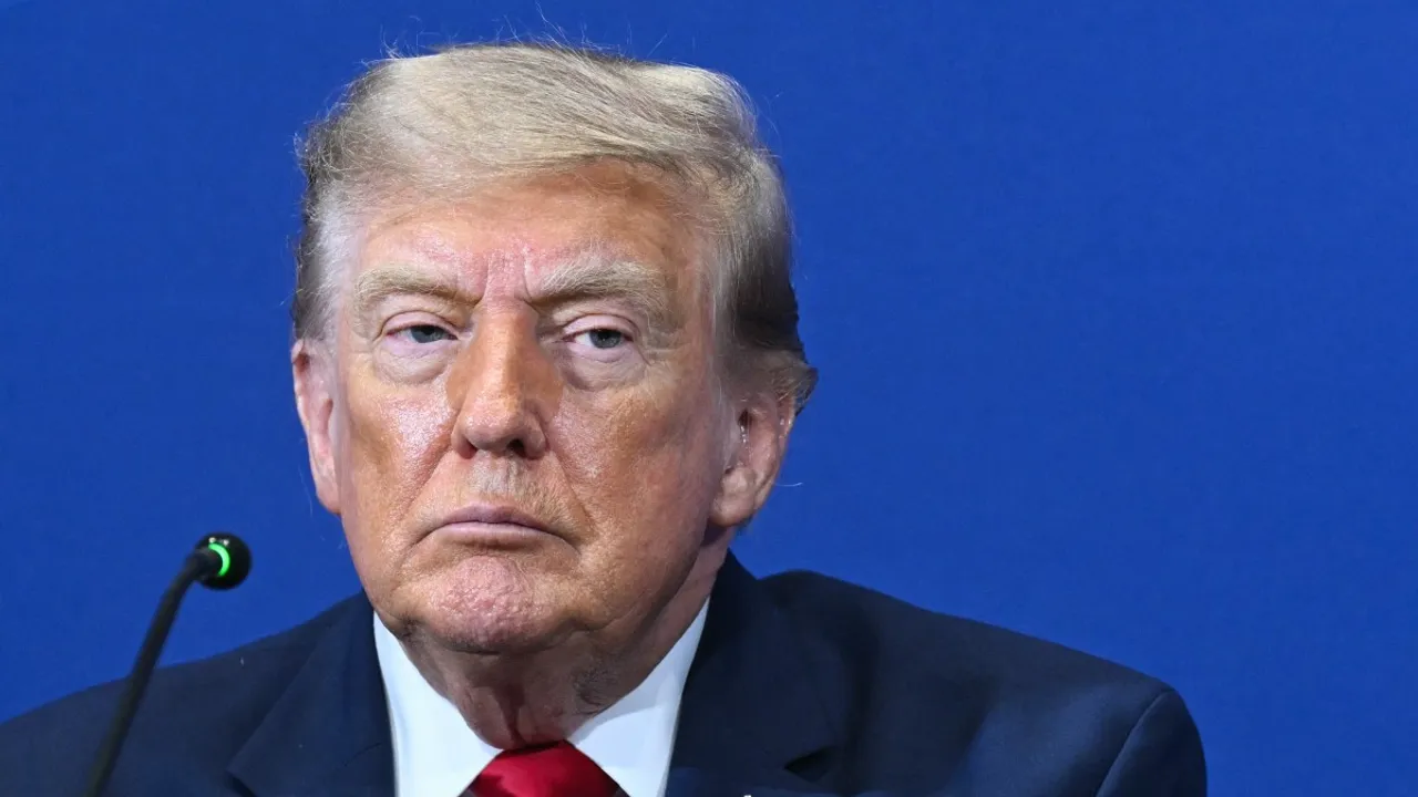 Primer plano del rostro de Donald Trump con expresión seria frente a un fondo azul y un micrófono.