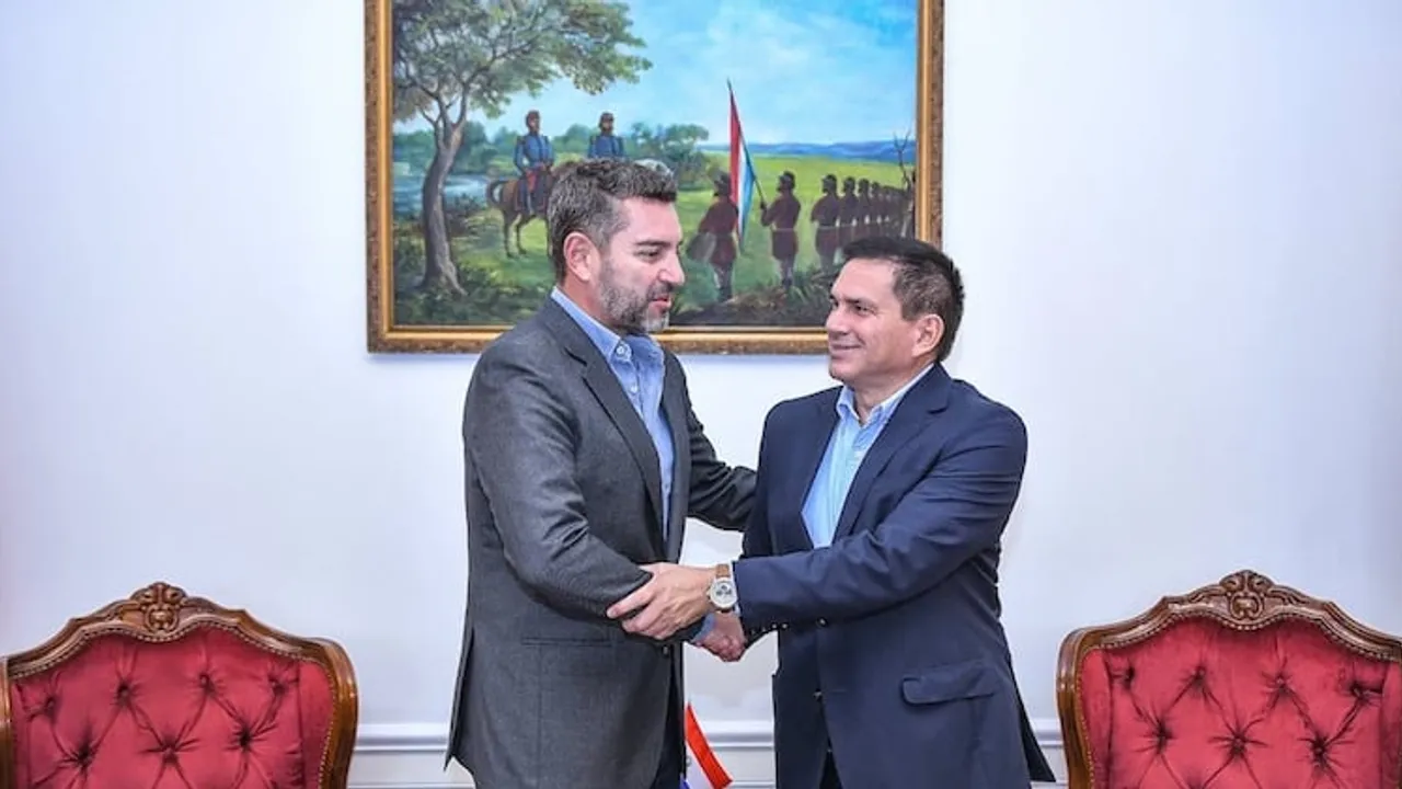 Pedro Alliana y Juan Carlos Baruja estrechan las manos de pie junto a dos sillones y un cuadro histórico en Asunción.