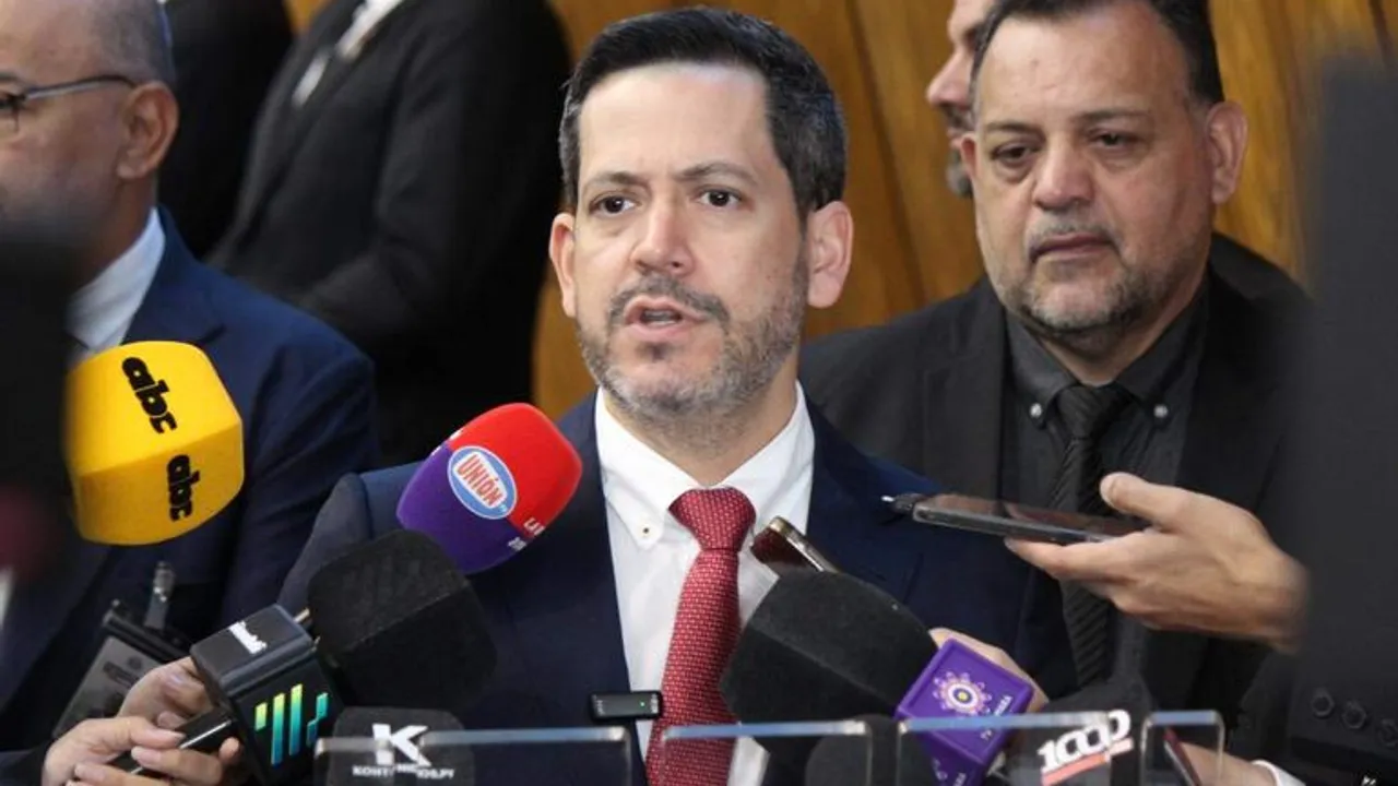 Hombre de traje y corbata habla rodeado de micrófonos y grabadores de prensa durante una rueda de medios.