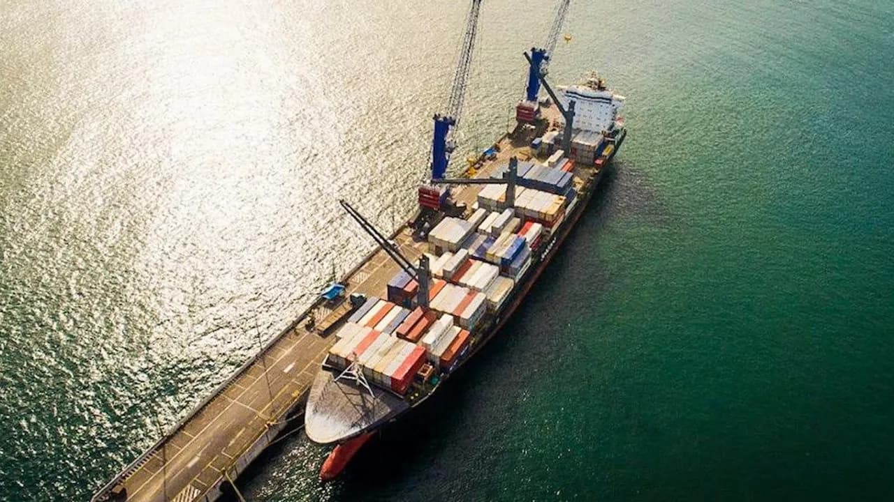 Barco carguero atracado en un muelle portuario con grúas de carga y contenedores metálicos sobre cubierta en el mar.