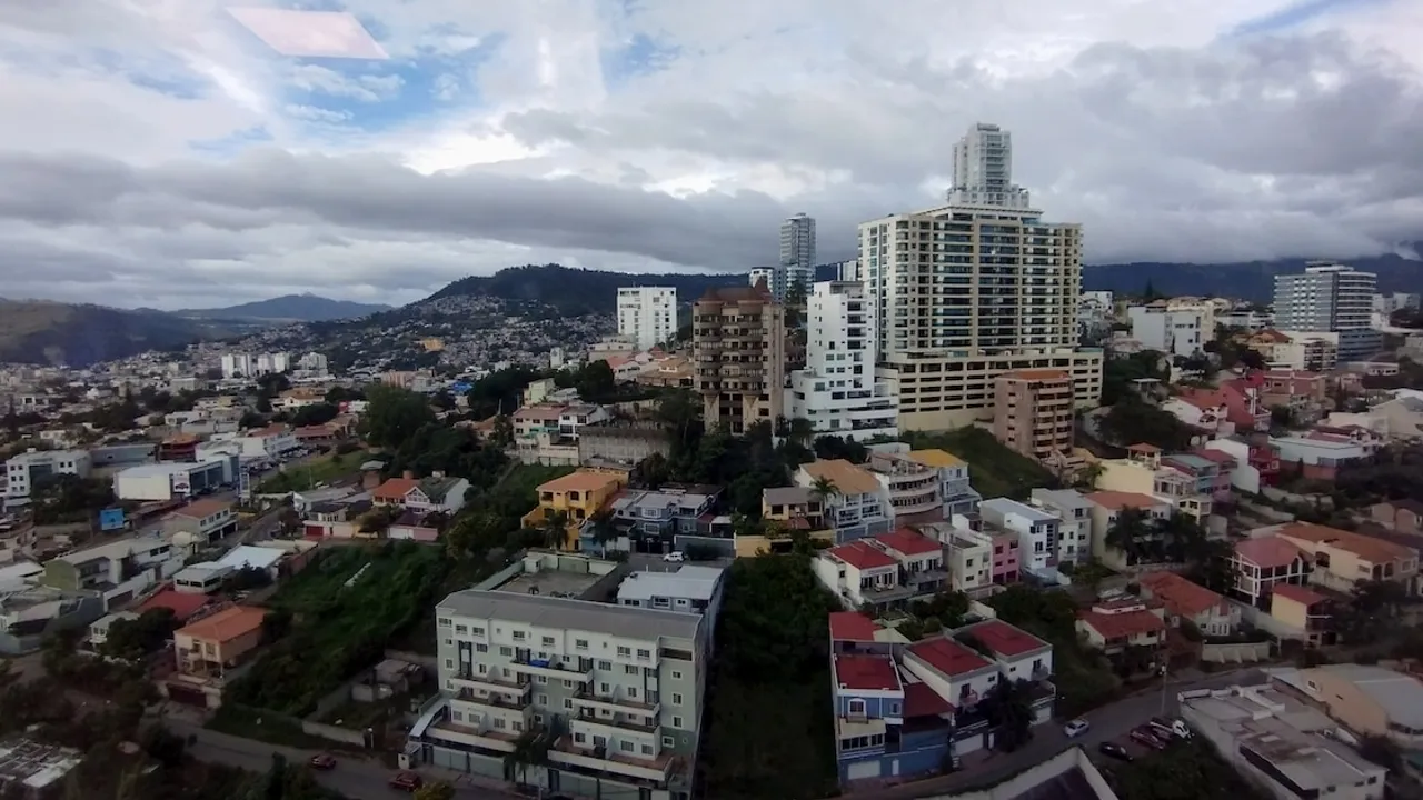 Vista panorámica urbana con edificios residenciales y casas sobre laderas bajo un cielo nublado.
