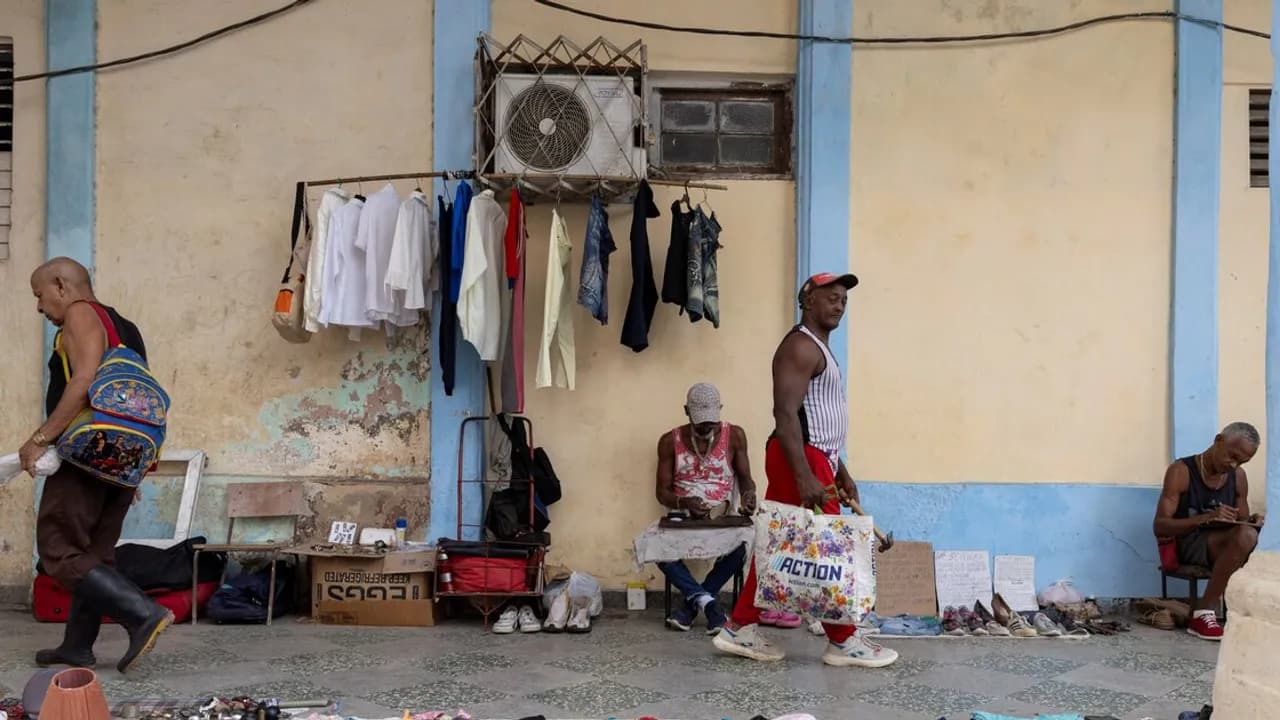 Personas sentadas en una acera ofrecen ropa colgada y calzados usados frente a una pared descascarada.