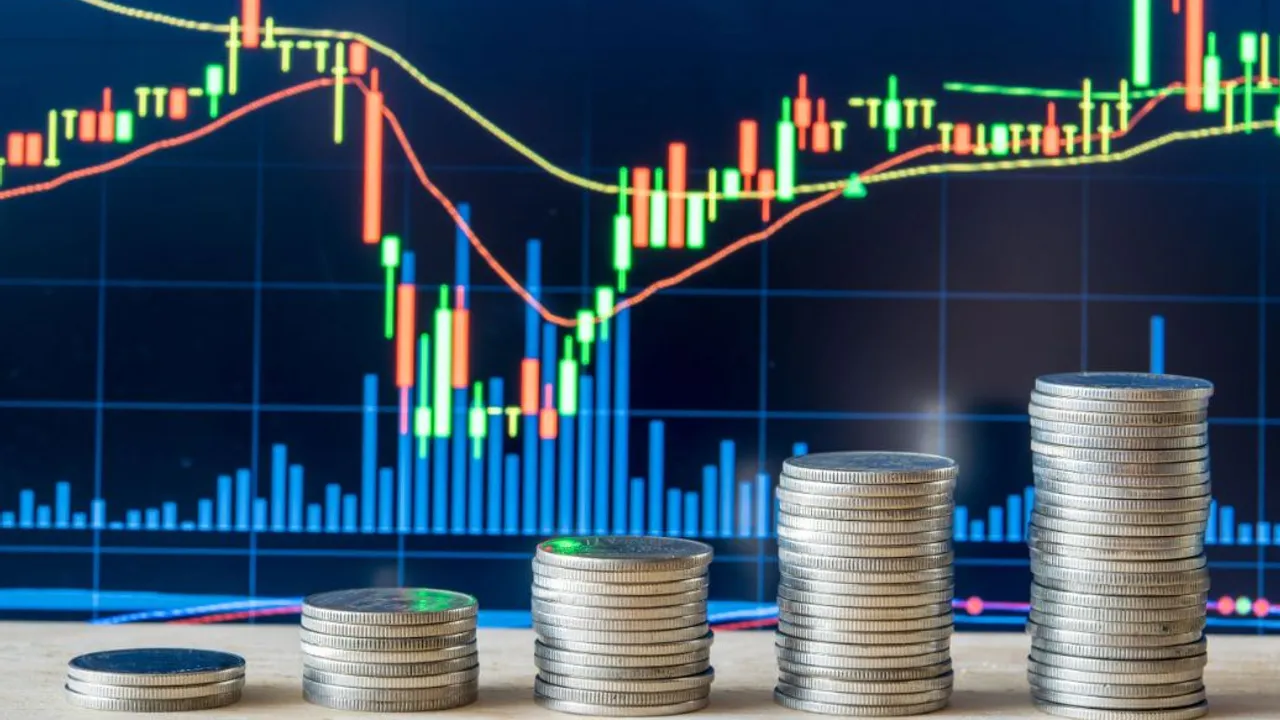 Columnas de monedas frente a una pantalla con gráficos bursátiles y curvas financieras.