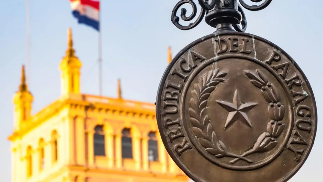 Escudo de metal de la República del Paraguay en primer plano frente a la fachada iluminada del Palacio de López.