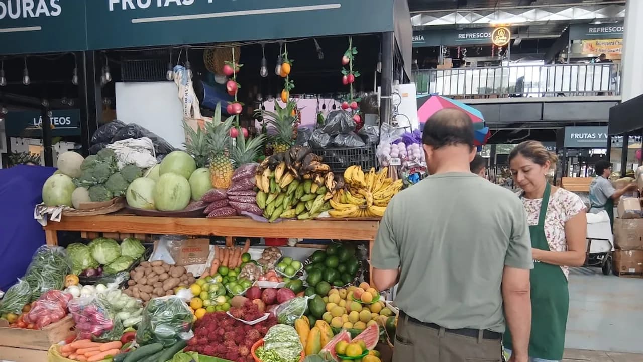 Un cliente observa un puesto repleto de frutas y verduras variadas en un mercado interior mientras la vendedora atiende.