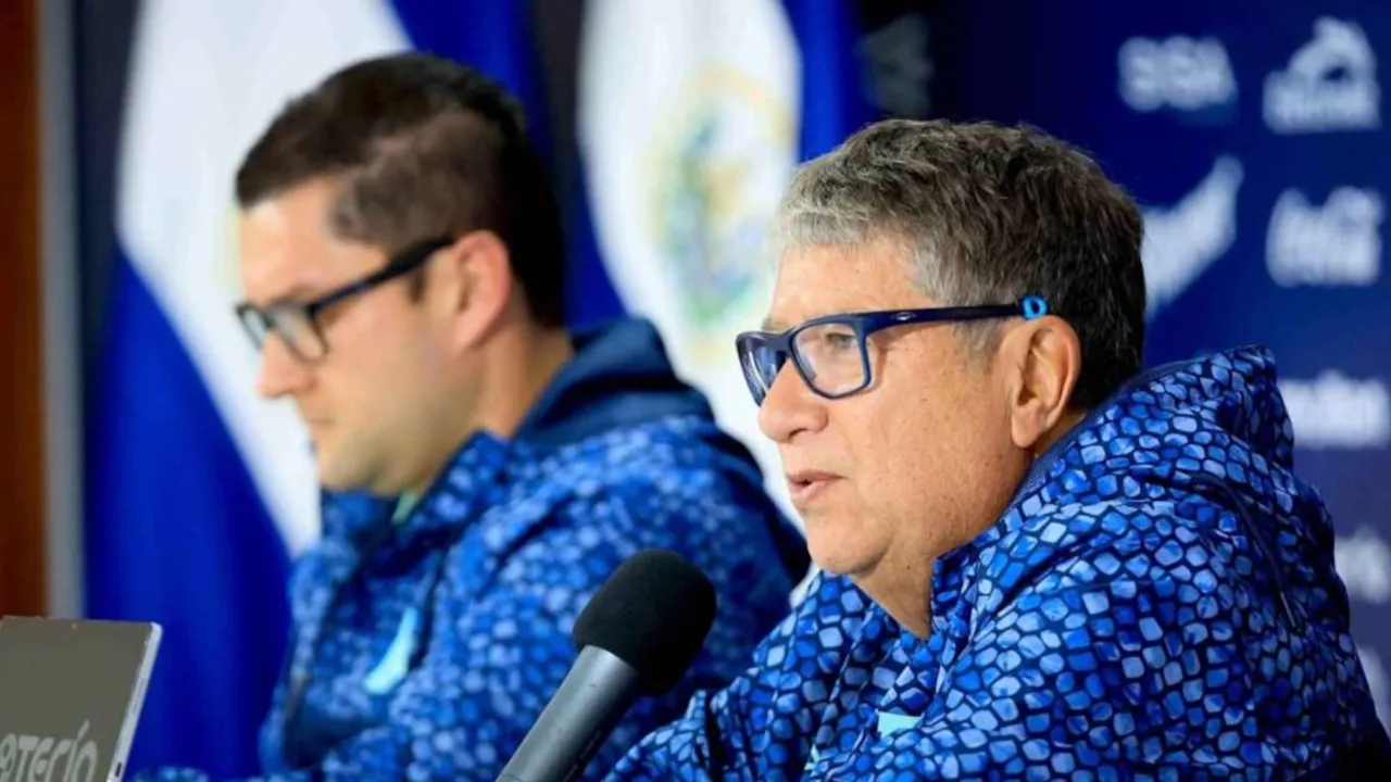 Hernán Darío Gómez con anteojos y campera azul habla frente a un micrófono en una rueda de prensa deportiva.