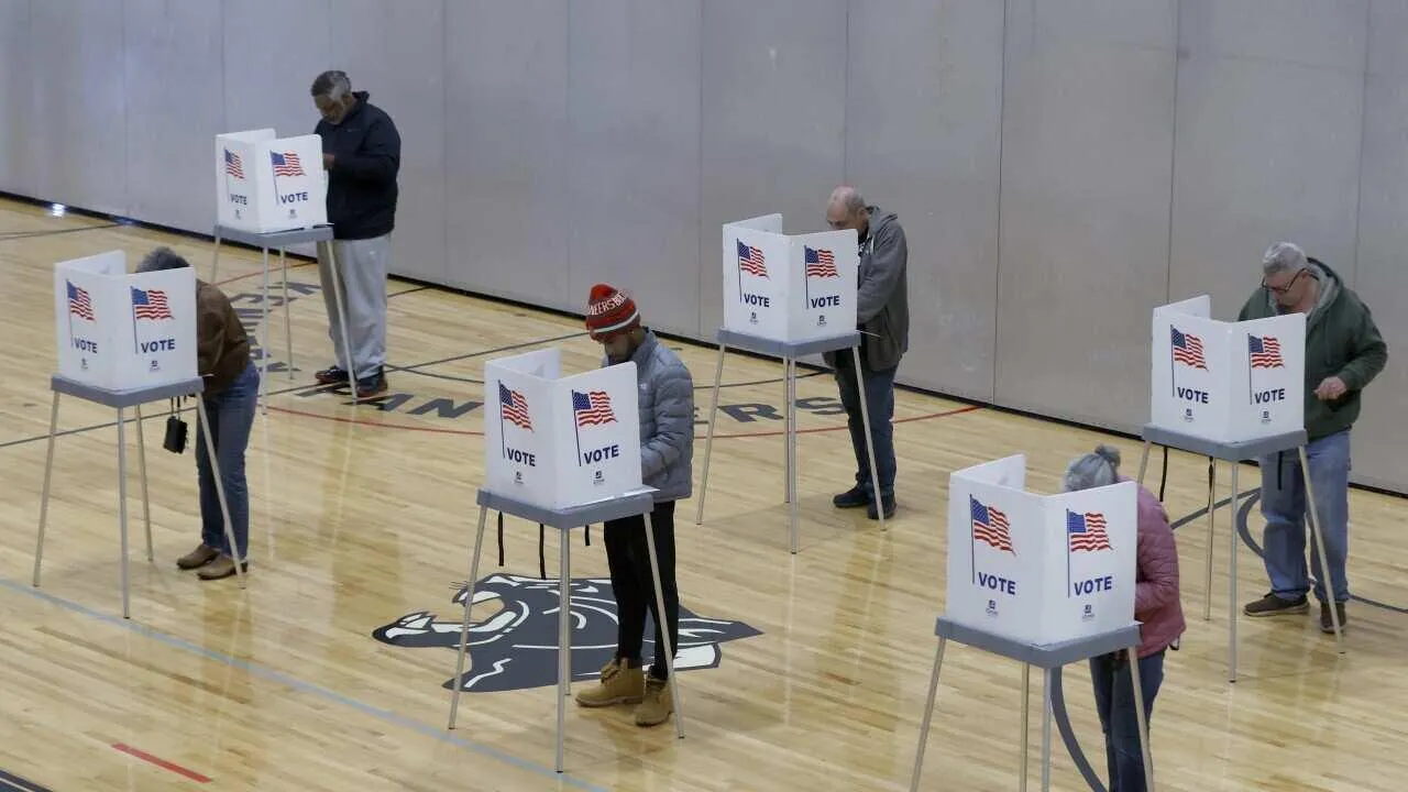 Ciudadanos emiten su voto de forma individual dentro de cabinas electorales con banderas en un gimnasio escolar.