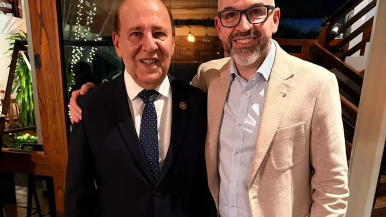 Dos hombres sonrientes con vestimenta formal se abrazan amistosamente en un ambiente interior iluminado.