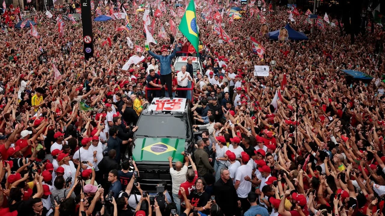 Multitud congregada en una avenida ondeando banderas rodea un vehículo durante un acto político en San Pablo.