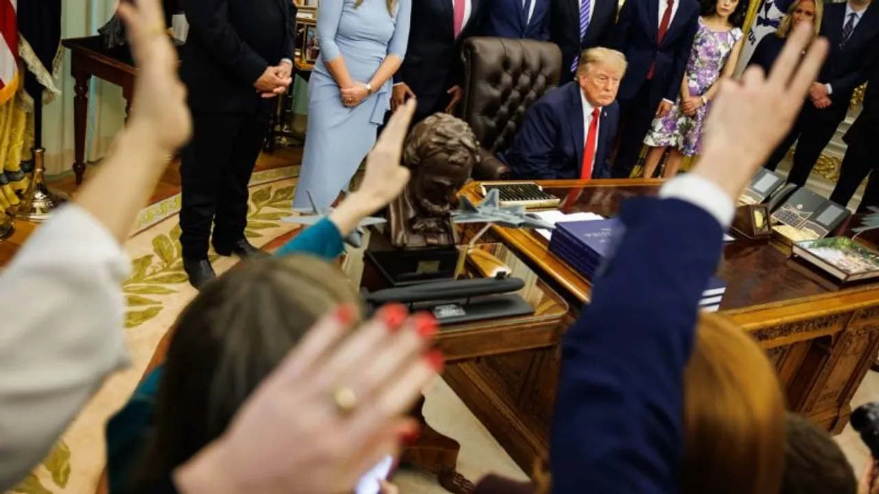 Donald Trump sentado tras su escritorio en el Despacho Oval rodeado de personas y periodistas levantando las manos