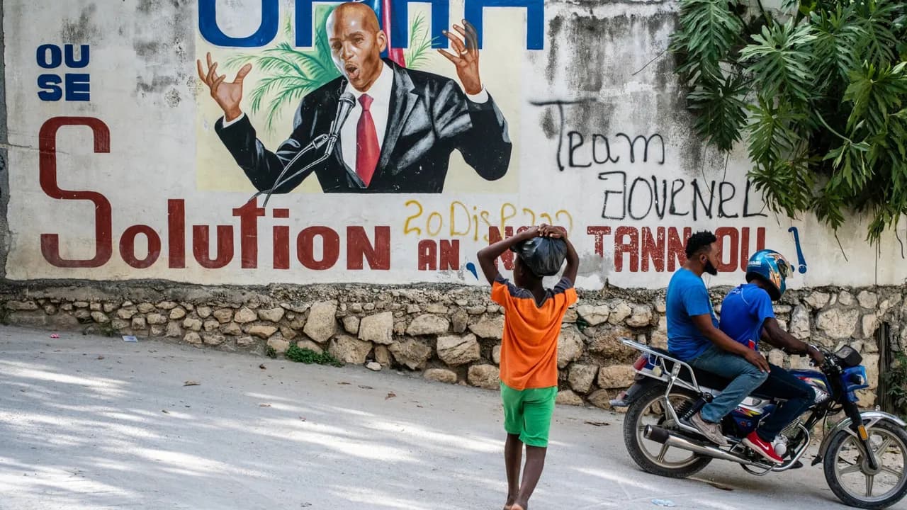 Mural con el rostro de Jovenel Moïse en una calle por donde circulan dos personas en motocicleta y un transeúnte.