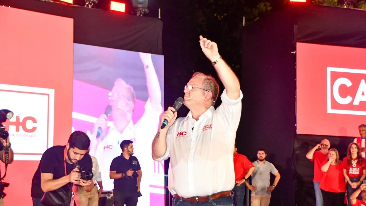 Hombre con camisa blanca y micrófono en mano habla en un escenario político nocturno señalando hacia arriba