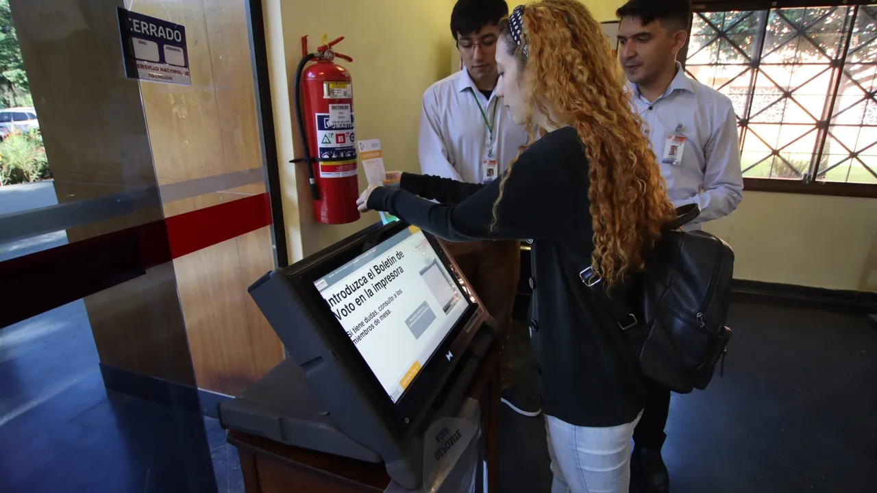 Una joven prueba una máquina de votación electrónica acompañada de dos funcionarios del área electoral.