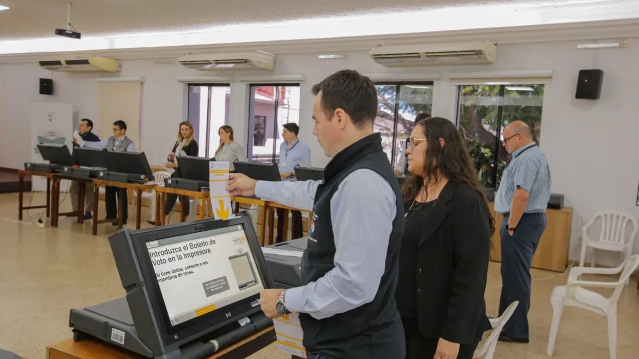 Personas realizando una demostración con una máquina de votación electrónica en un salón iluminado.