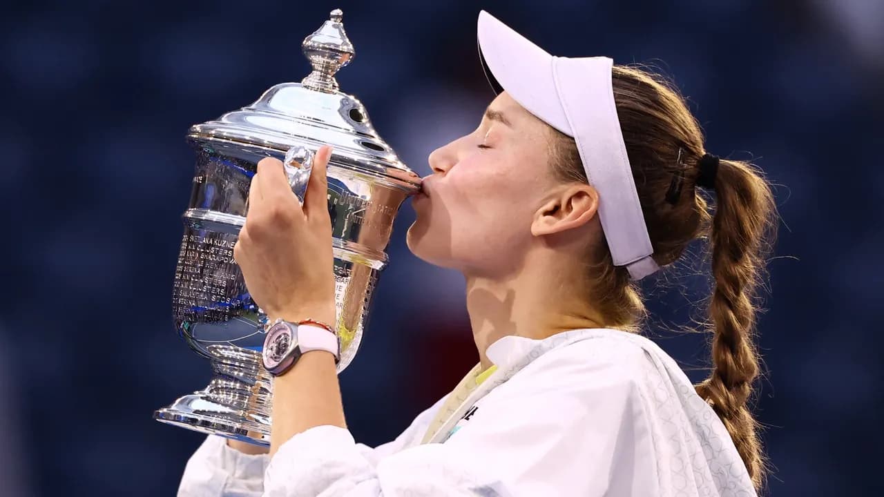 Tenista femenina con visera blanca sostiene y besa un gran trofeo plateado en una pista de tenis.