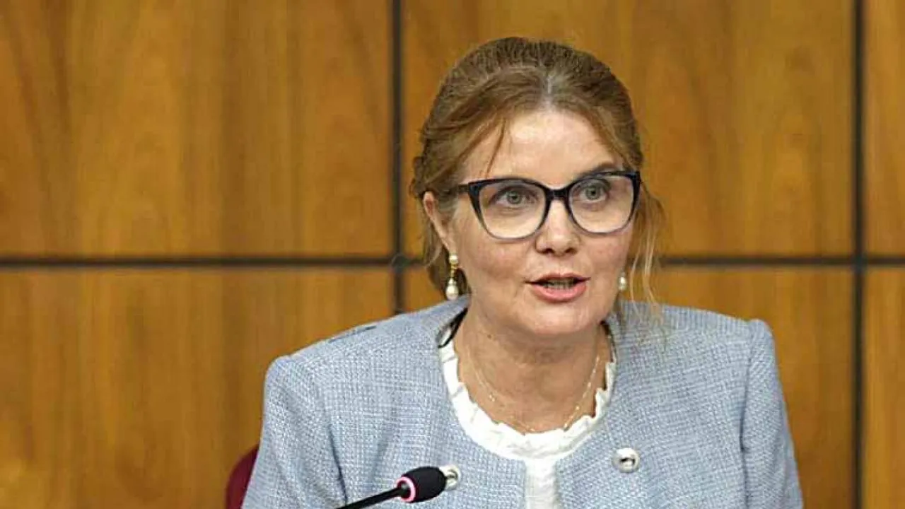 Mujer con anteojos y saco claro habla frente a un micrófono en una mesa con panel de madera.