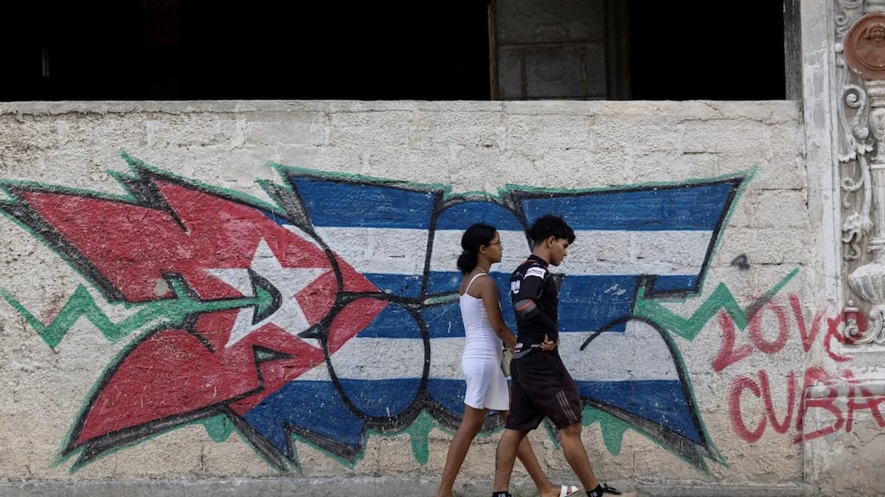 Dos personas jóvenes caminan junto a un muro que muestra un grafiti con los colores y la estrella de la bandera de Cuba.