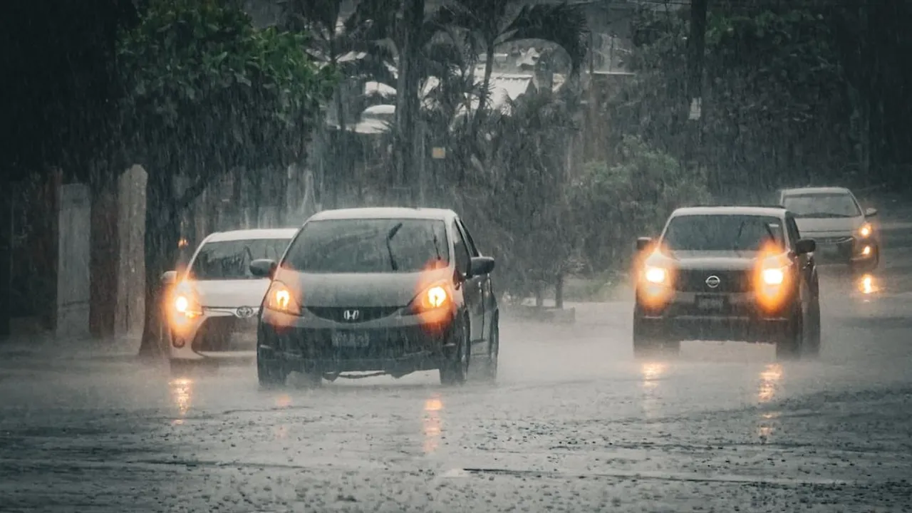 Vehículos circulando con luces encendidas bajo una intensa lluvia sobre una avenida anegada.