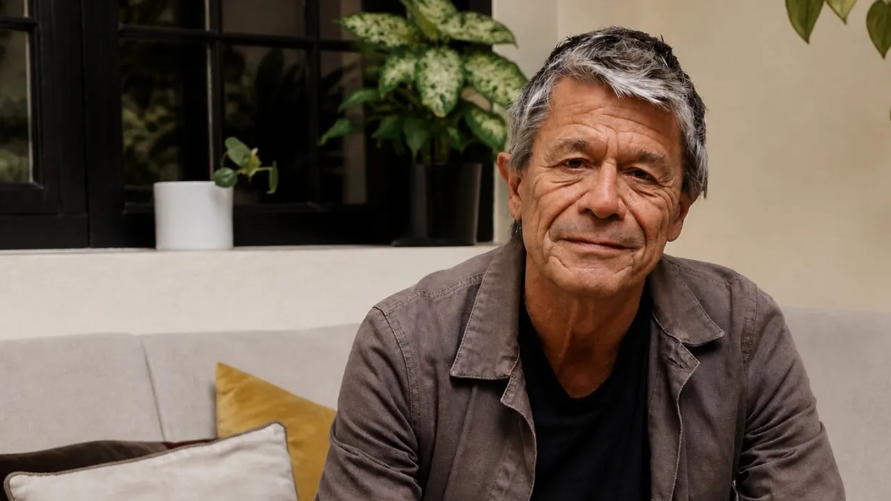 El escritor Emmanuel Carrère posa sentado en un sillón interior con plantas de fondo vistiendo una chaqueta marrón.