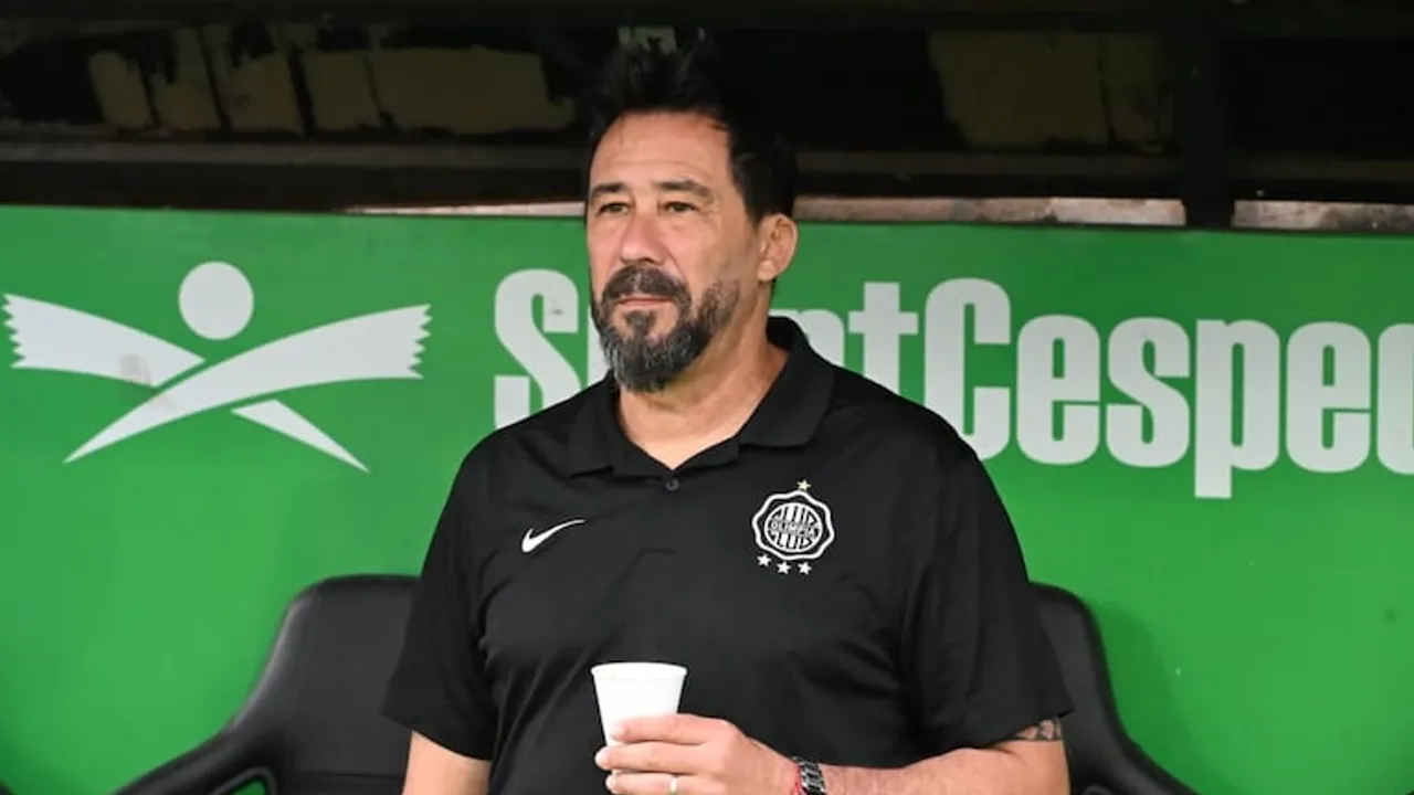 Pablo Vitamina Sánchez en la banca técnica con indumentaria oscura del Club Olimpia y un vaso en mano.