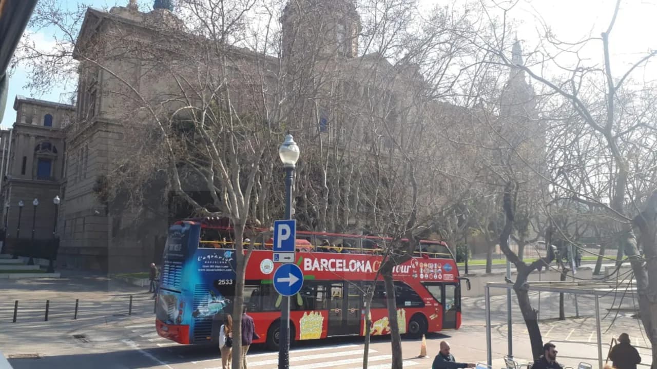 Bus turístico de dos pisos color rojo transitando por una avenida frente a un edificio antiguo en Barcelona.