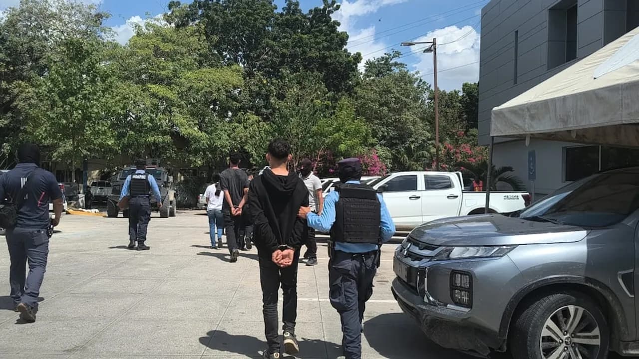 Agentes policiales custodian y trasladan a personas esposadas en un predio con patrullas y vehículos.