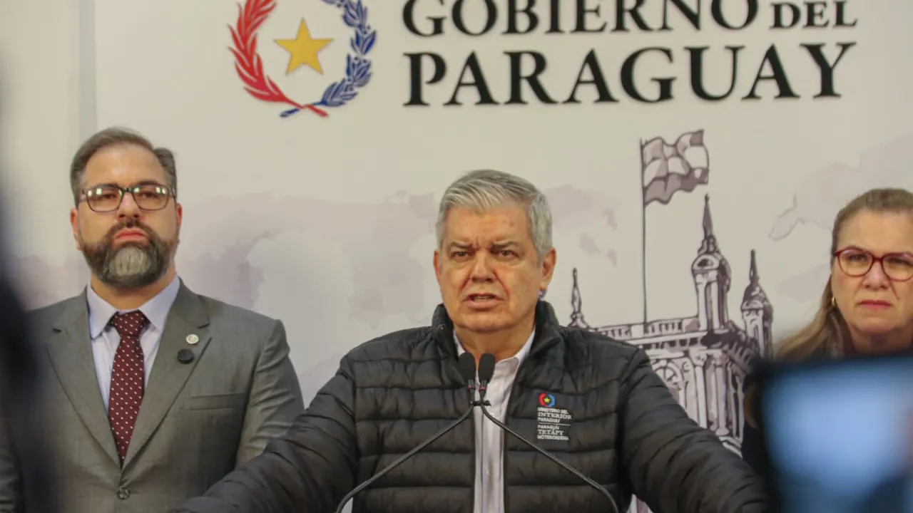 Enrique Riera habla frente a micrófonos con dos acompañantes detrás y el logo de Gobierno del Paraguay.