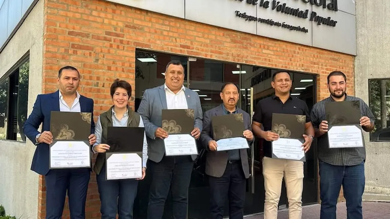 Seis personas posan sonrientes sosteniendo carpetas con diplomas frente a un edificio electoral con fachada de ladrillo.
