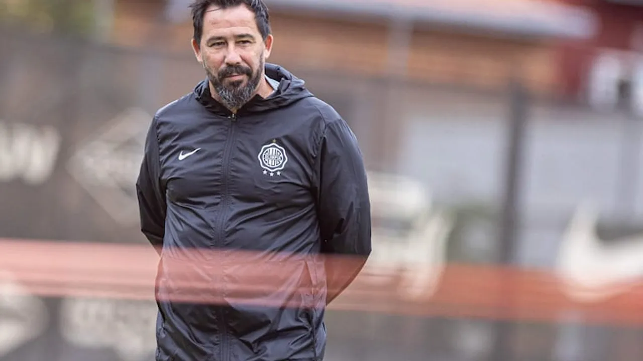 Hombre con barba y campera deportiva negra con el escudo de Olimpia parado en un campo de entrenamiento.