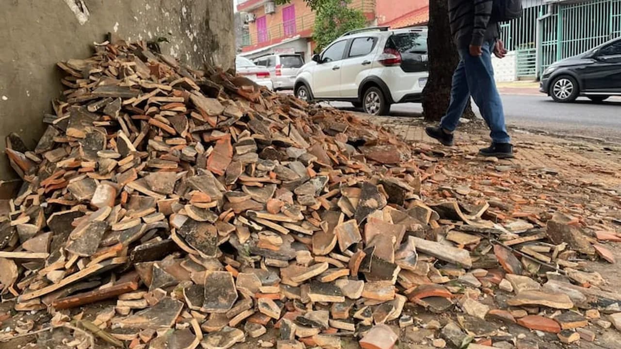 Gran montículo de tejas rotas y escombros sobre una vereda de la ciudad con un peatón caminando al lado.