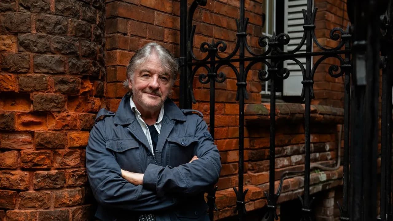 El escritor John Connolly posa de brazos cruzados con campera azul frente a una pared de ladrillos y reja negra.