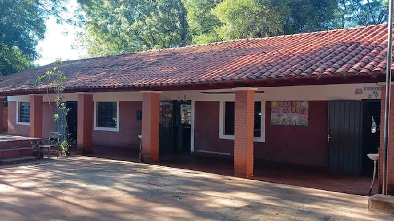 Fachada de una escuela rural con techo de tejas rojas, pilares de ladrillo visto y patio de tierra.