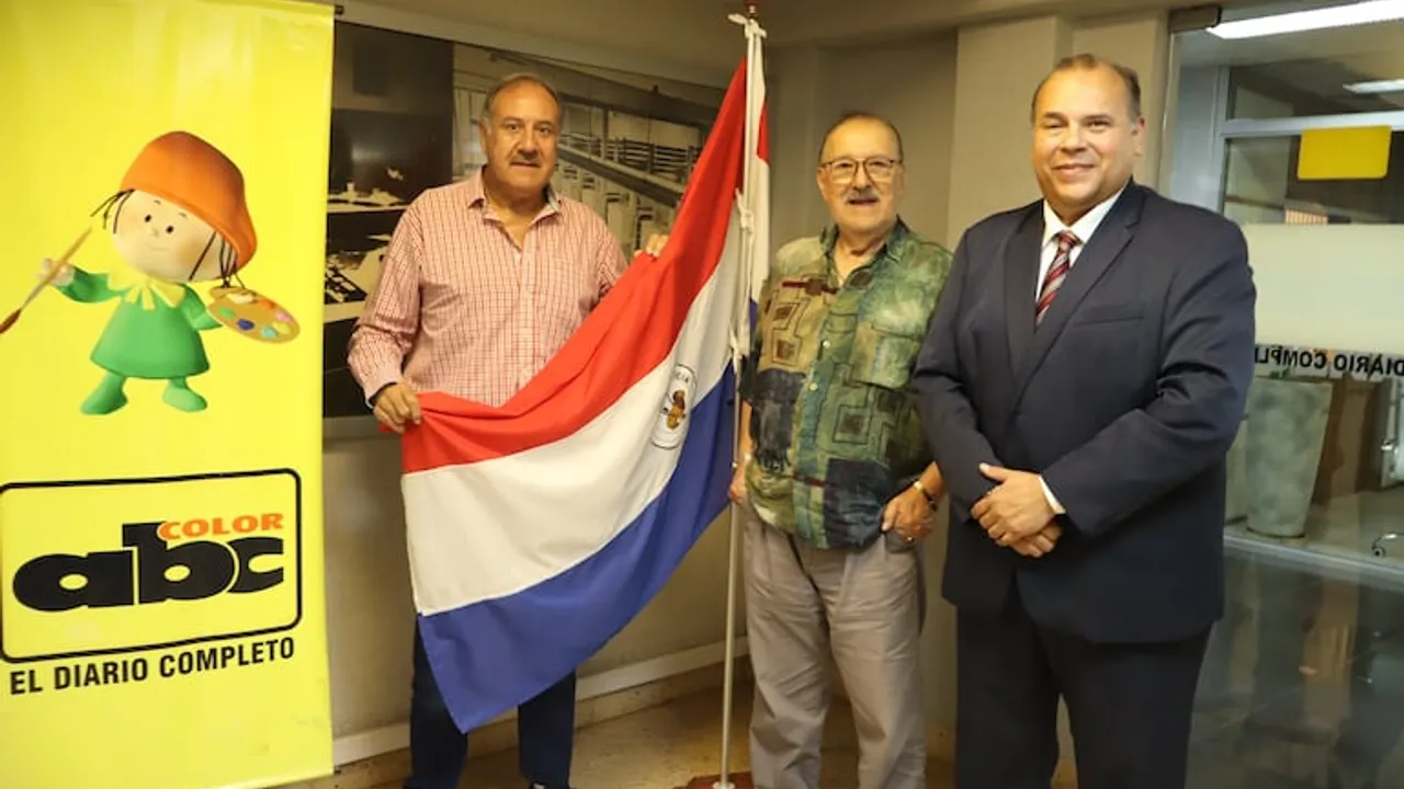 Tres hombres de pie junto a una bandera paraguaya y un cartel informativo dentro de una sala u oficina.