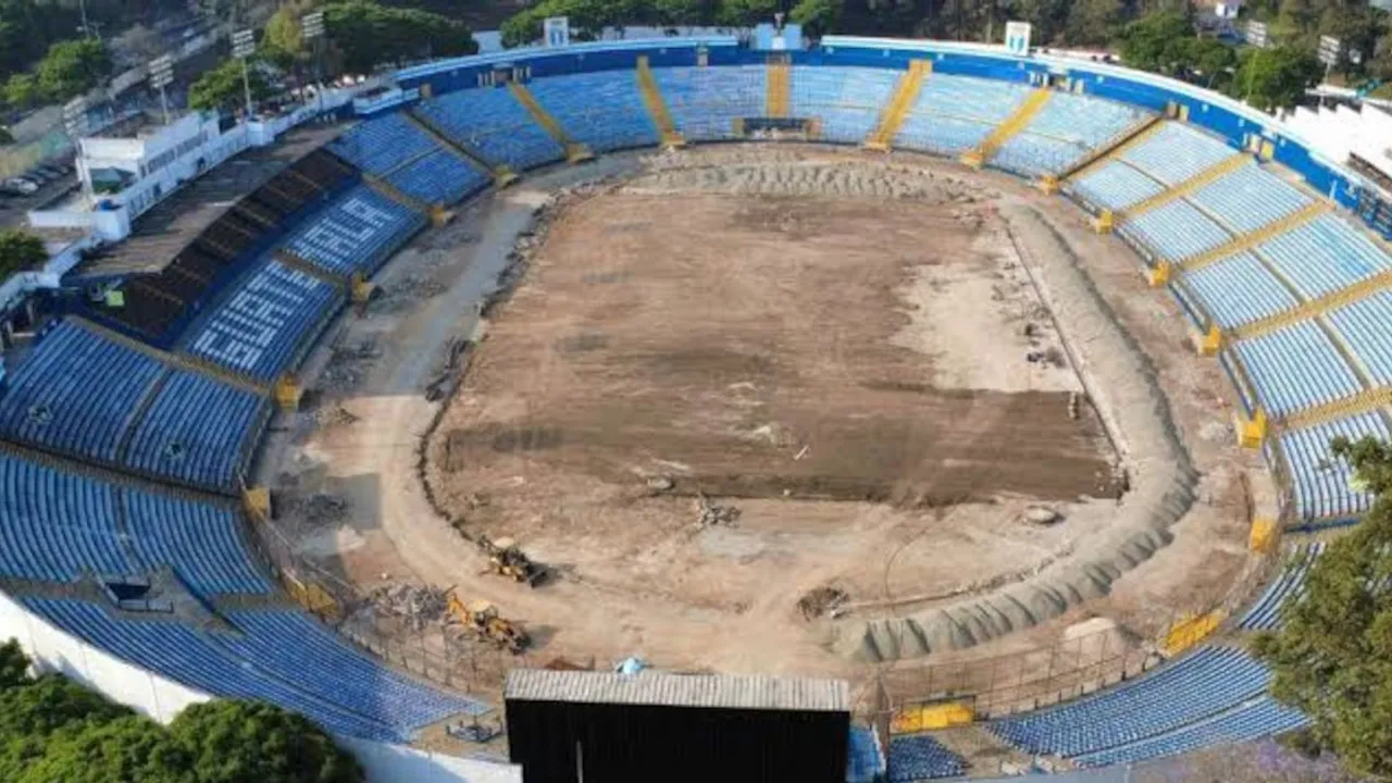 Vista aérea del campo de tierra y graderías azules de un estadio deportivo desmantelado durante obras.