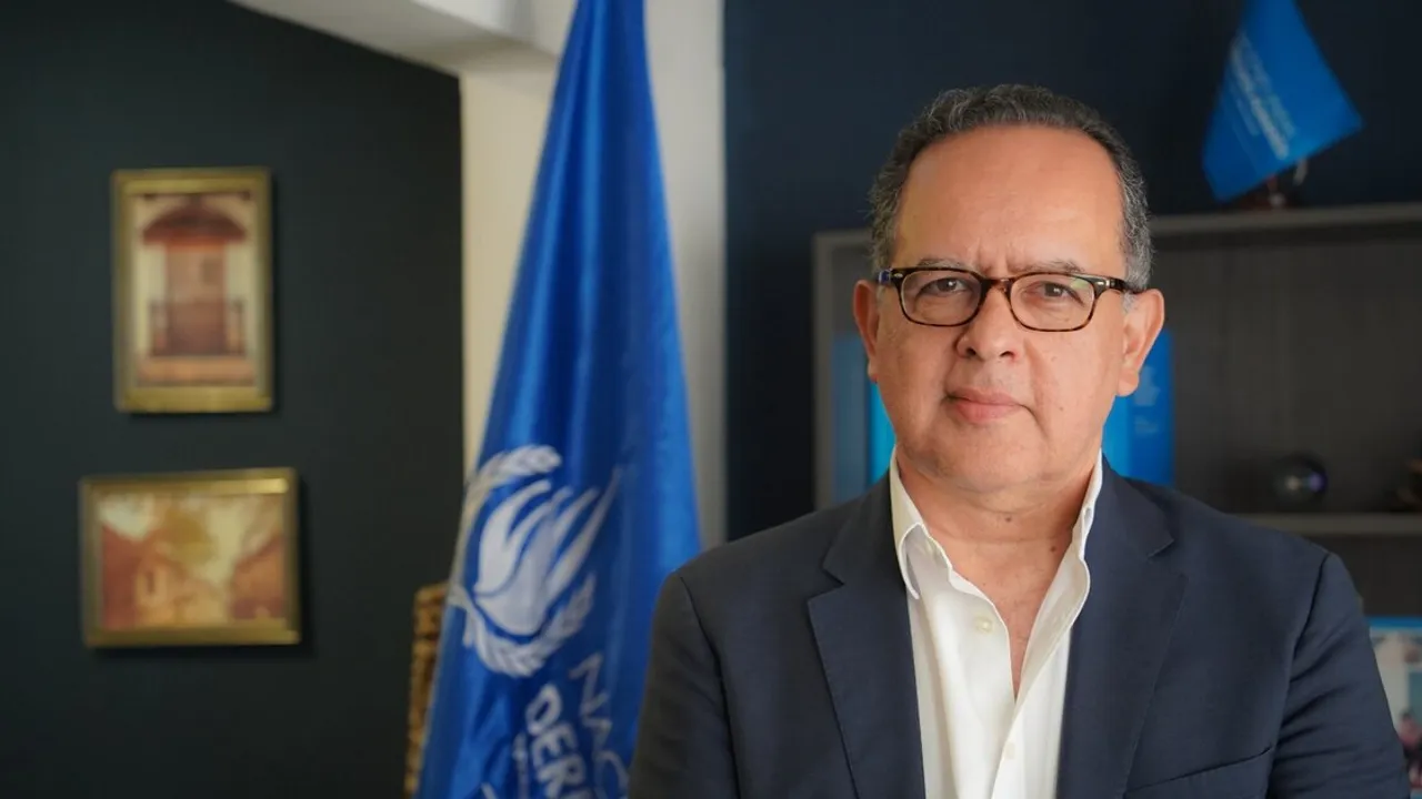 Juan Carlos Monge con lentes y traje oscuro junto a la bandera azul de Naciones Unidas en una oficina.
