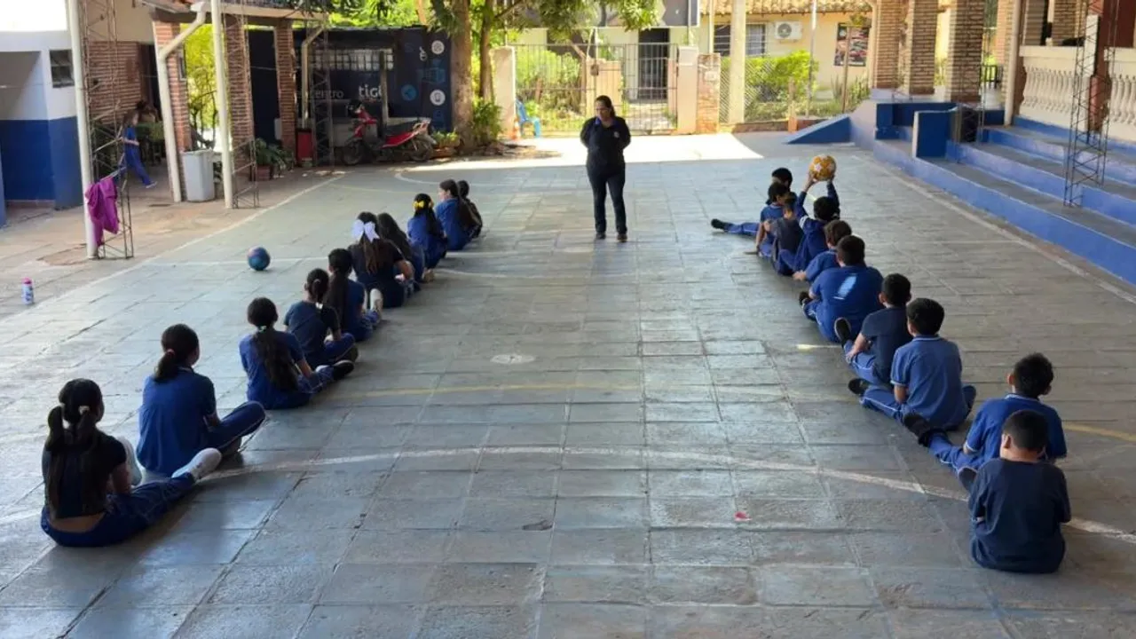 Estudiantes sentados en filas en el patio escolar observan una pelota durante una clase al aire libre.