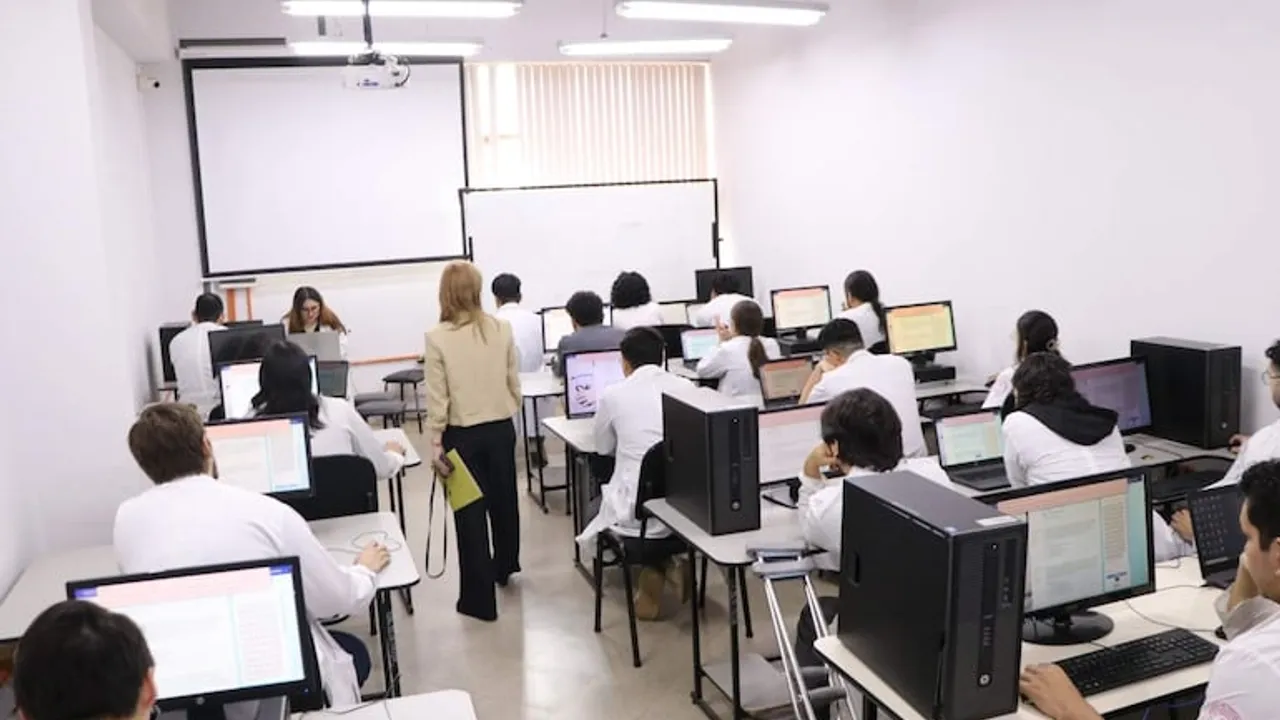 Estudiantes con batas blancas rinden una prueba frente a computadoras en una sala informática con una evaluadora