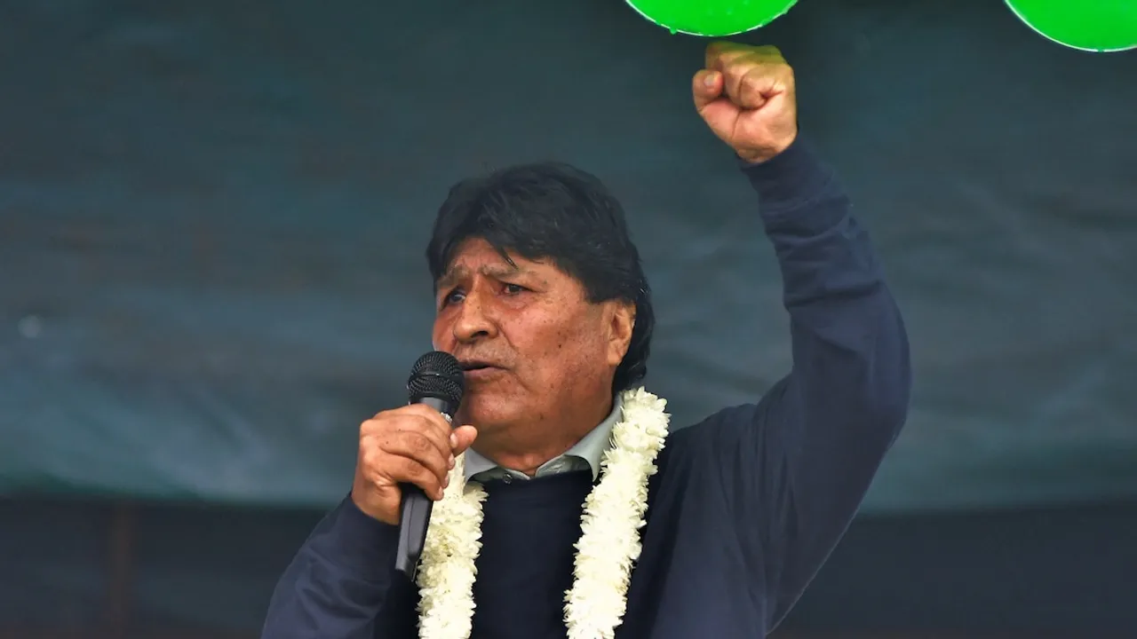 Evo Morales habla por micrófono con un collar de flores blancas y el puño izquierdo levantado en alto.
