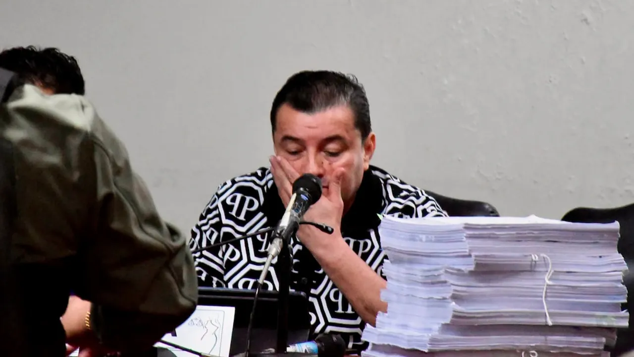 Hombre con camisa estampada sentado detrás de un micrófono y una pila de documentos en un estrado judicial.