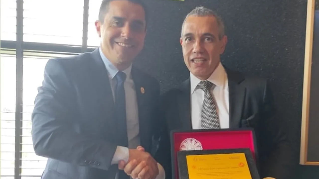 Dos hombres en traje se estrechan la mano mientras sostienen una placa de reconocimiento en una oficina.