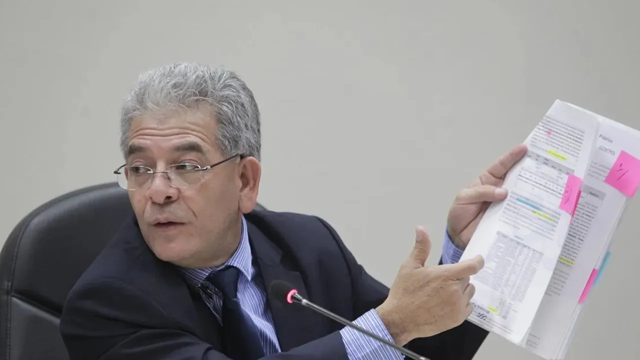 Hombre con lentes y traje oscuro muestra un expediente con notas adhesivas frente a un micrófono en una sala.